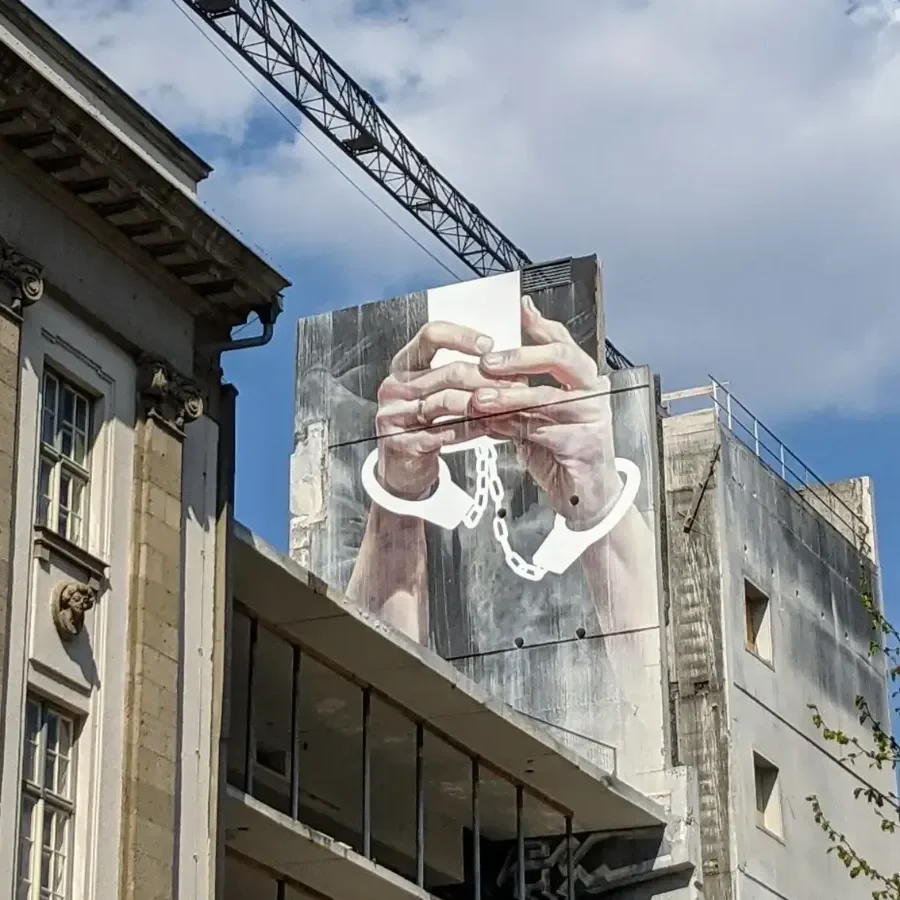 Wandbild Cell Phone von Innerfields am früheren Parkhaus an der Karl-Marx-Straße in Berlin-Neukölln: zwei Hände in Handschellen halten ein Smartphone, darüber ein Baukran