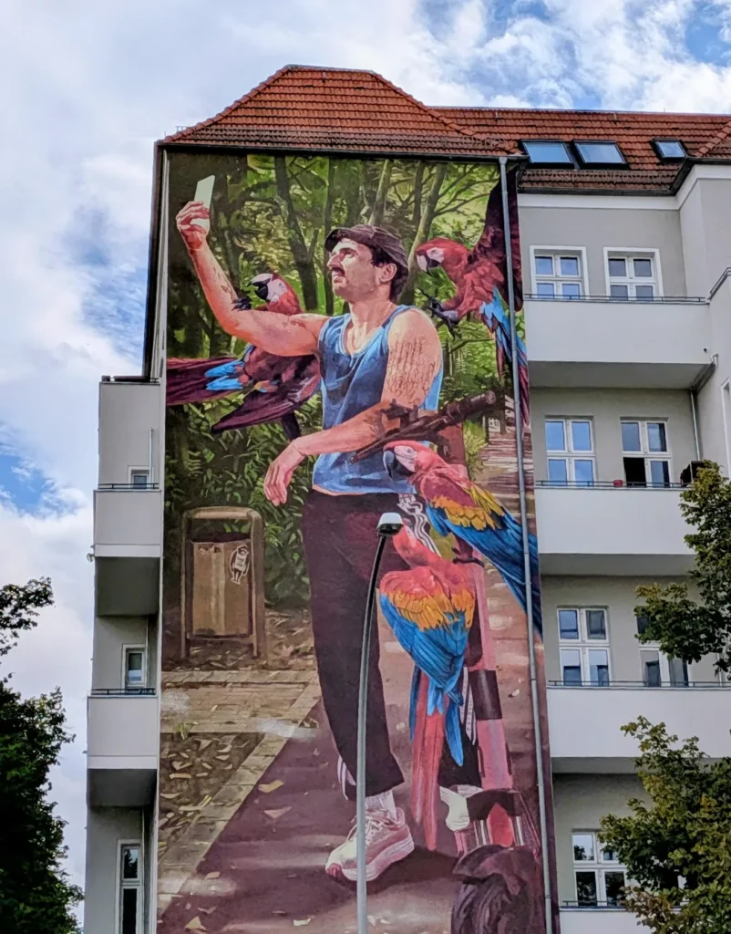 Wandbild Papageienmann von Innerfields an der Schivelbeiner Straße in Berlin-Prenzlauer Berg: Mann im Tanktop macht ein Selfie, um ihn sitzen vier Aras
