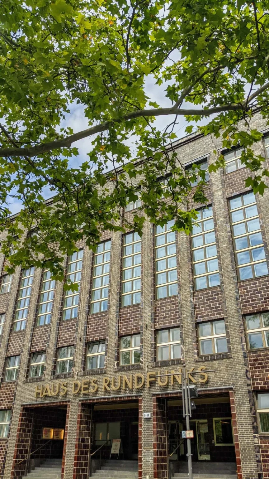 Dunkle Klinkerfassade des Hauses des Rundfunks von schräg unten, goldene Buchstaben über dem Eingang, im oberen Bilddrittel Platanenäste