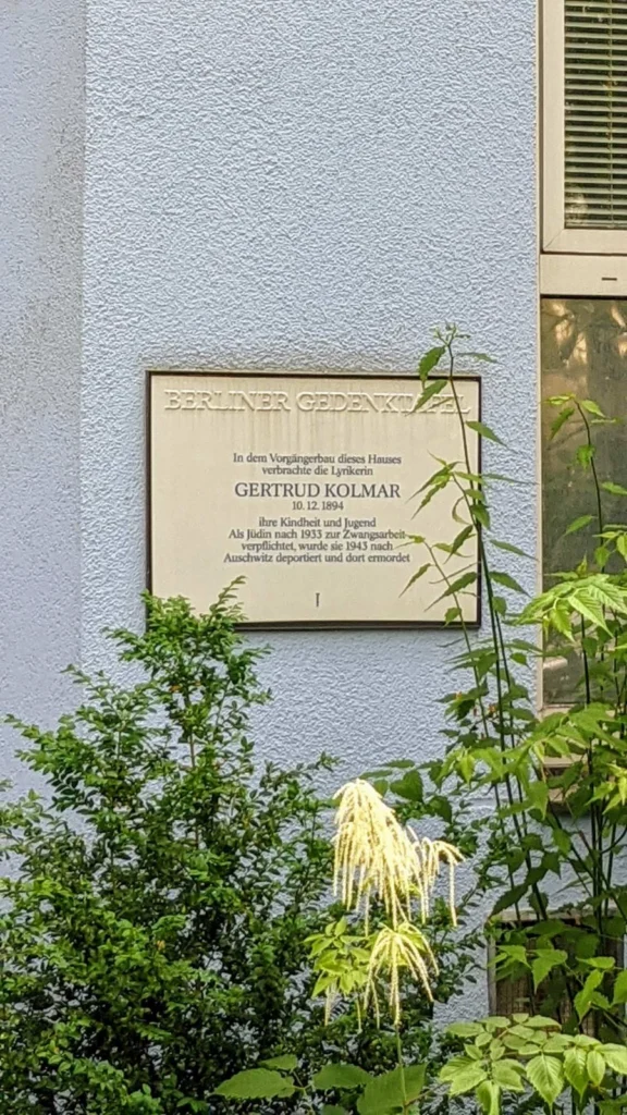 Berliner Gedenktafel aus Porzellan für die Lyrikerin Gertrud Kolmar an einer hellblauen Putzfassade, davor Sträucher und Stauden