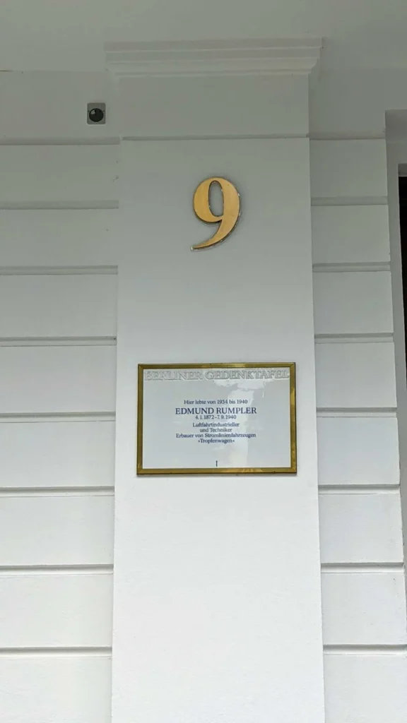 Berliner Gedenktafel mit goldfarbenem Rahmen für Edmund Rumpler an einem weißen Hauspfeiler, darüber die goldene Hausnummer 9