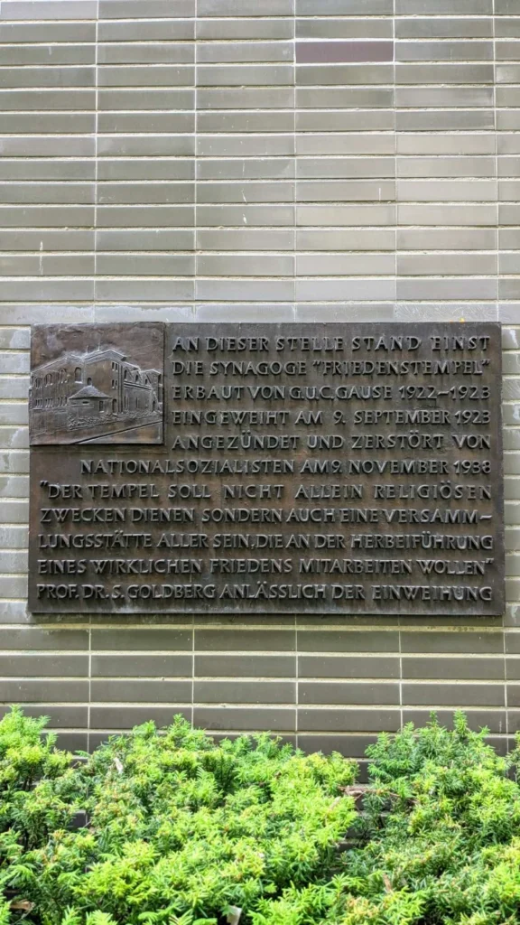 Bronzetafel mit dem Relief eines Synagogengebäudes an einer hellen Riemchenwand, unten eine Eibenhecke, sie erinnert an die Synagoge Friedenstempel in Halensee