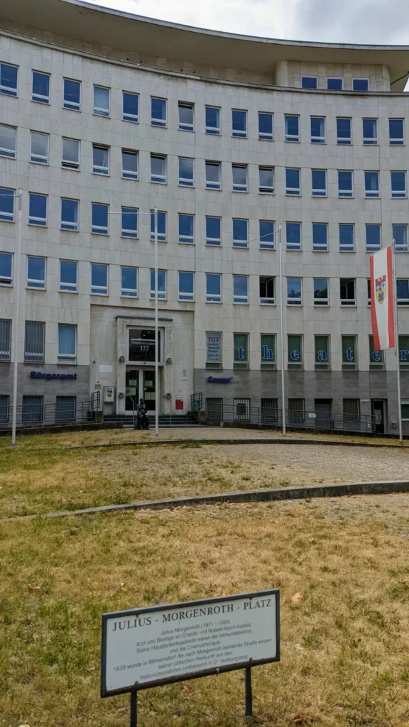 Geschwungenes Verwaltungsgebäude mit Natursteinfassade am Hohenzollerndamm 177, davor ein Erläuterungsschild zum Julius-Morgenroth-Platz auf trockenem Rasen und eine bronzene Sitzfigur