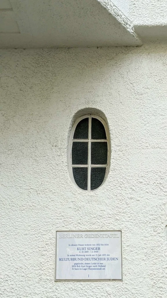 Berliner Gedenktafel für Kurt Singer und den Kulturbund Deutscher Juden an einer weißen Rauputzfassade unter einem ovalen Sprossenfenster