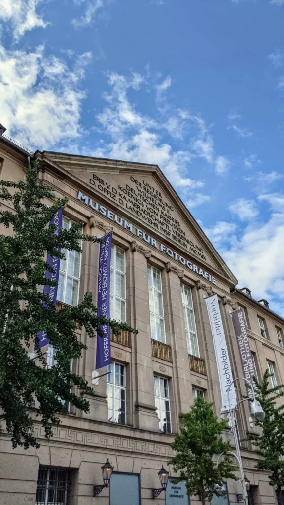 Neoklassizistische Sandsteinfassade des Museums für Fotografie mit Dreiecksgiebel und Ausstellungsbannern vor blauem Himmel mit Wolken
