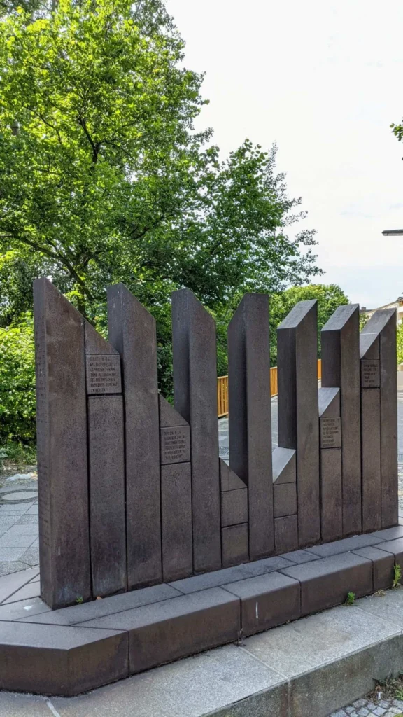 Mahnmal aus dunkelbraunen, gestaffelten Stahlpfeilern mit oben abgeschrägten Enden auf einem Sockel, in die Pfeiler sind kleine Inschriftfelder eingelassen, dahinter Bäume und ein gelbes Brückengeländer