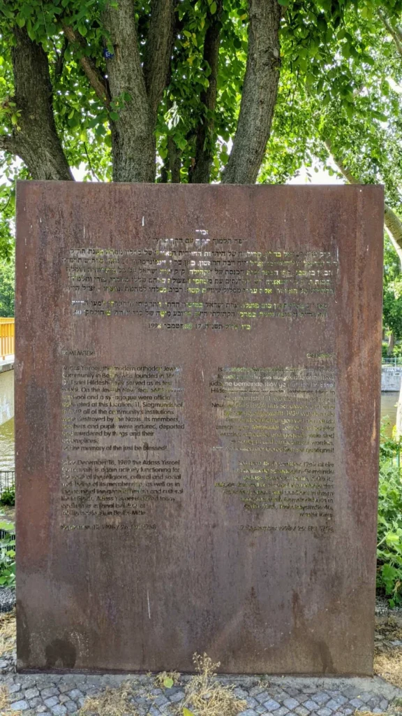 Aufrechte Stahlplatte mit dreisprachiger Inschrift, oben hebräisch, darunter links englisch und rechts deutsch, unter einem mehrstämmigen Baum am Spreeufer