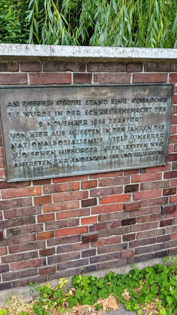 Grün patinierte Bronzetafel an einer roten Ziegelmauer mit Betonabdeckung, darüber Weidenzweige, sie erinnert an die zerstörte Synagoge in der Levetzowstraße