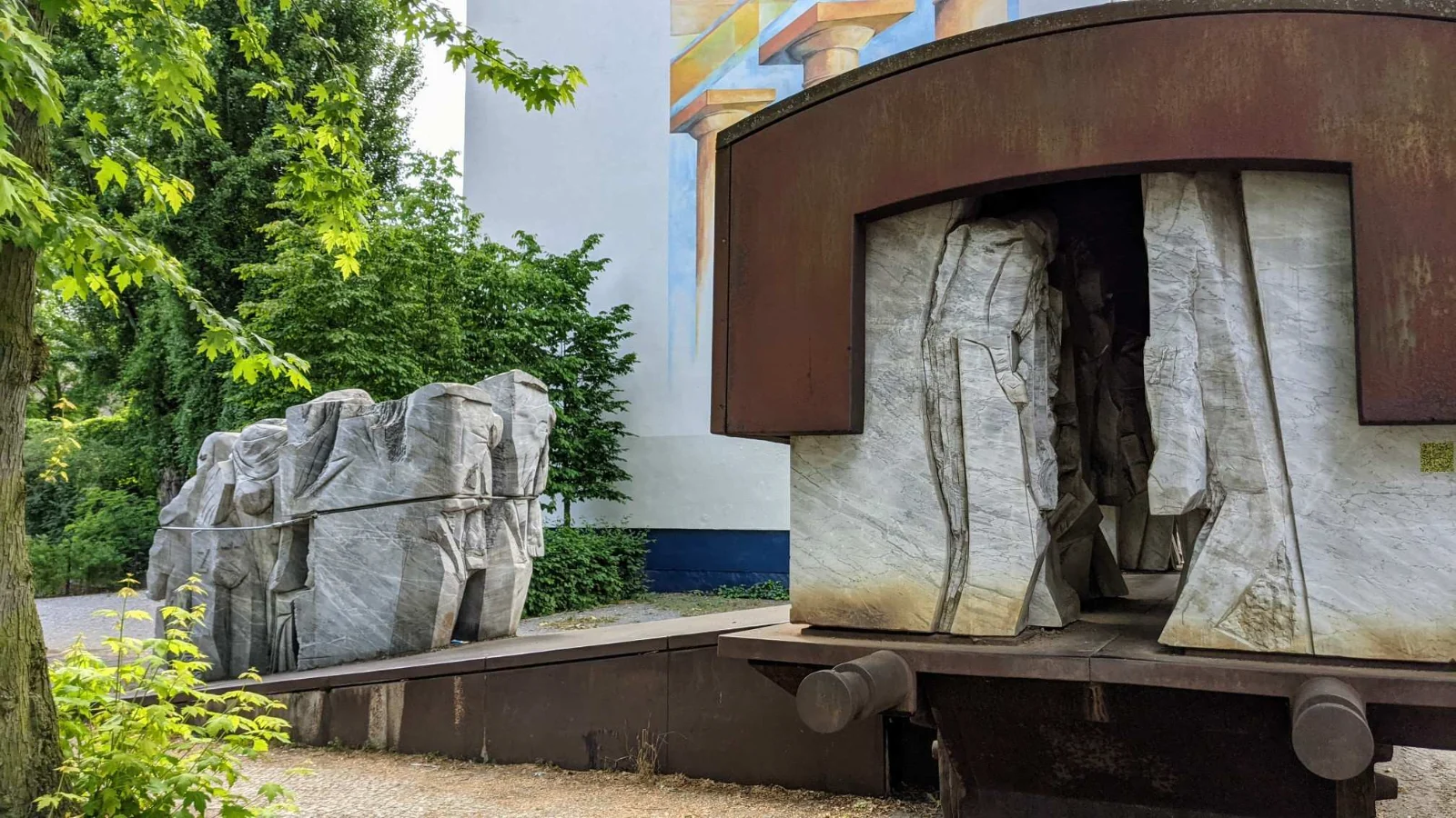 Mahnmal Levetzowstraße: rechts ein stählerner Waggonrahmen mit Puffern und grob behauenen Marmorfiguren, links eine Rampe mit einer zusammengeschnürten Figurengruppe aus Marmor, dahinter eine Brandwand und Bäume