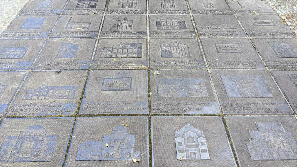 Bodenfeld aus Eisenplatten, jede mit dem Relief einer Berliner Synagoge und einer Textzeile zu Adresse, Sitzplätzen, Baujahr und Schicksal, dazwischen Laub
