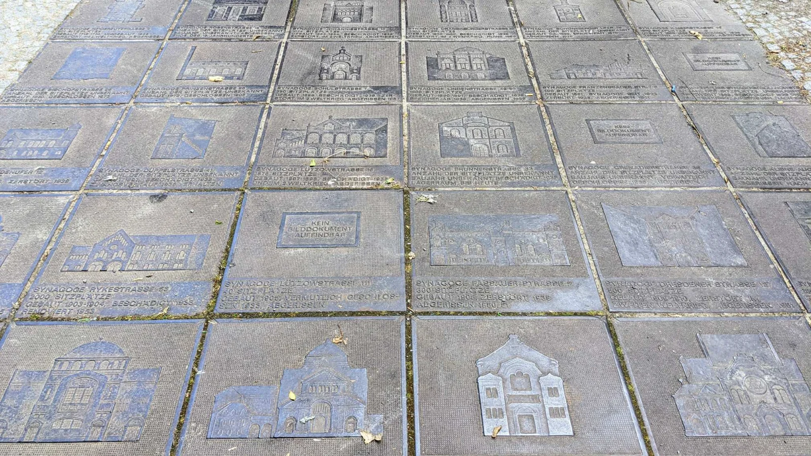 Bodenfeld aus Eisenplatten, jede mit dem Relief einer Berliner Synagoge und einer Textzeile zu Adresse, Sitzplätzen, Baujahr und Schicksal, dazwischen Laub