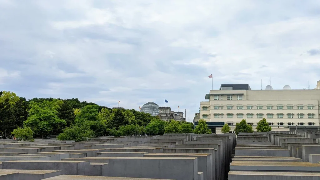 Feld grauer Betonstelen unter bedecktem Himmel, dahinter eine Baumreihe, eine Glaskuppel und ein Gebäude mit US-Flagge