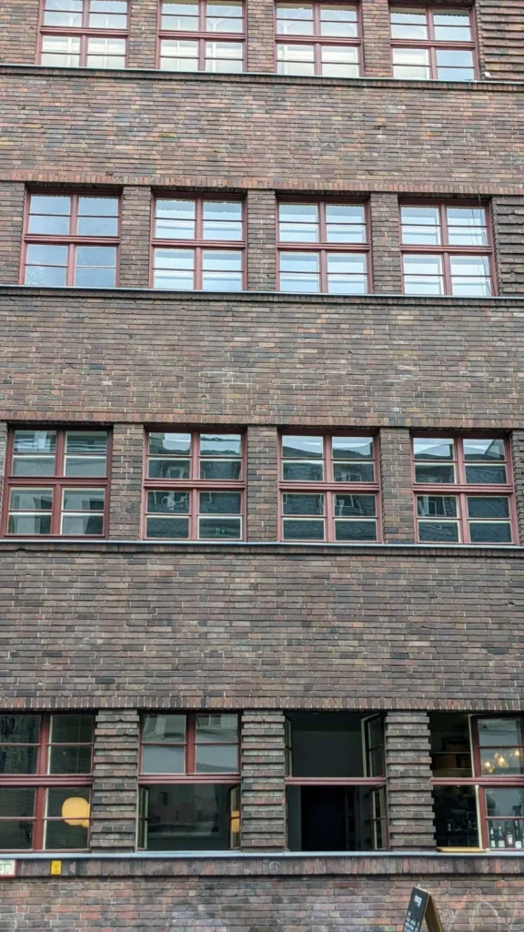 Dunkelrote Klinkerfassade mit vier Fensterreihen und roten Sprossenfenstern, im Erdgeschoss der Blick in ein Lokal, unten rechts ein Aufsteller