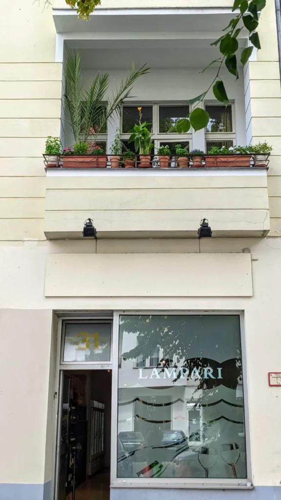 Hellgelbe Hausfassade mit bepflanztem Balkon, darunter ein Ladenfenster mit dem Schriftzug Lampari und aufgemalten Weingläsern, die Ladentür steht offen
