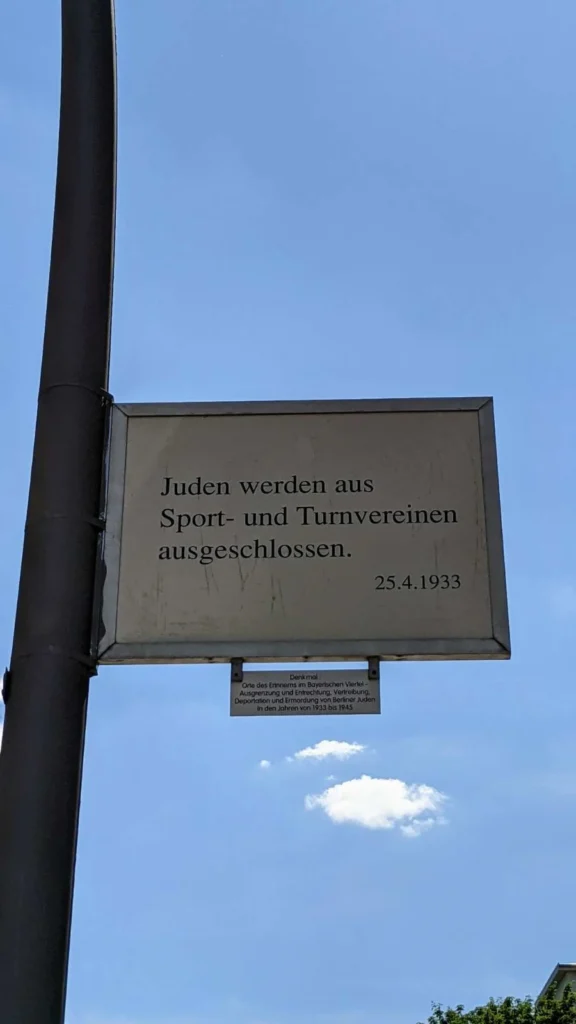 Schild des Denkmals Orte des Erinnerns im Bayerischen Viertel an einem Laternenmast, Text zum Ausschluss jüdischer Menschen aus Sport- und Turnvereinen am 25. April 1933