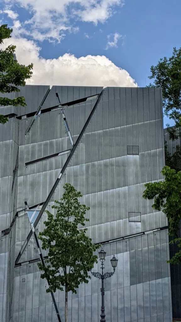 Fensterlose Zinkfassade des Libeskind-Baus des Jüdischen Museums mit schräg eingeschnittenen Fensterbändern, davor ein junger Baum