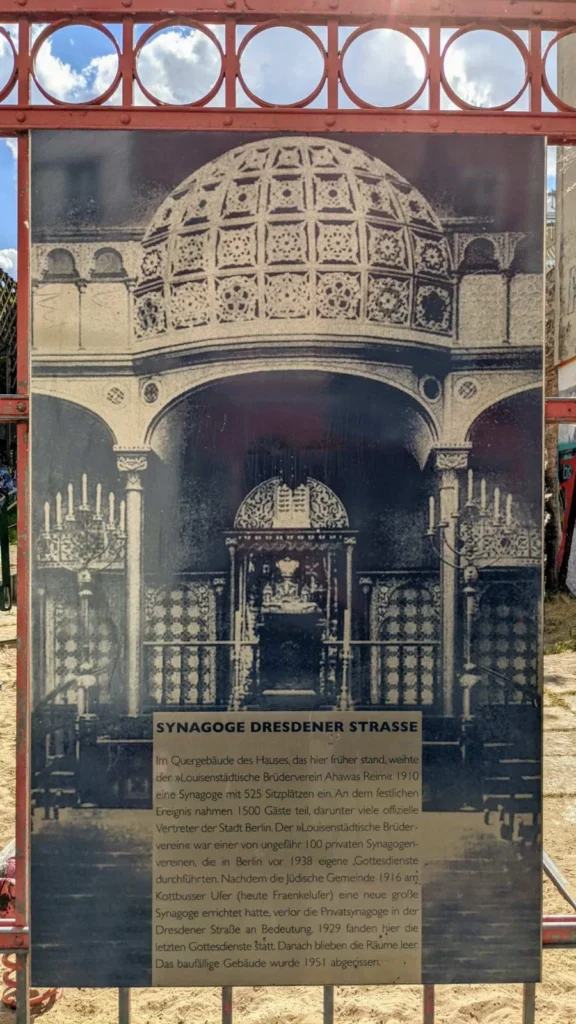 Infotafel Synagoge Dresdener Straße in rotem Stahlrahmen am Zaun eines Spielplatzes, oben ein historisches Foto des Innenraums, darunter der Text