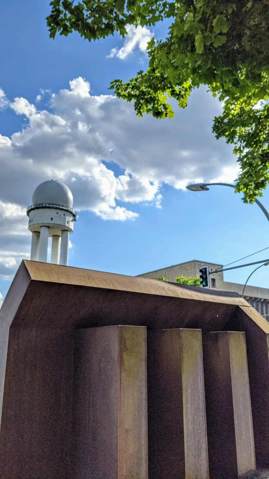 Rostbraune Stahlskulptur in Hausform mit Satteldach als Mahnmal für das KZ Columbia-Haus am Columbiadamm, dahinter ein weißer Radarturm