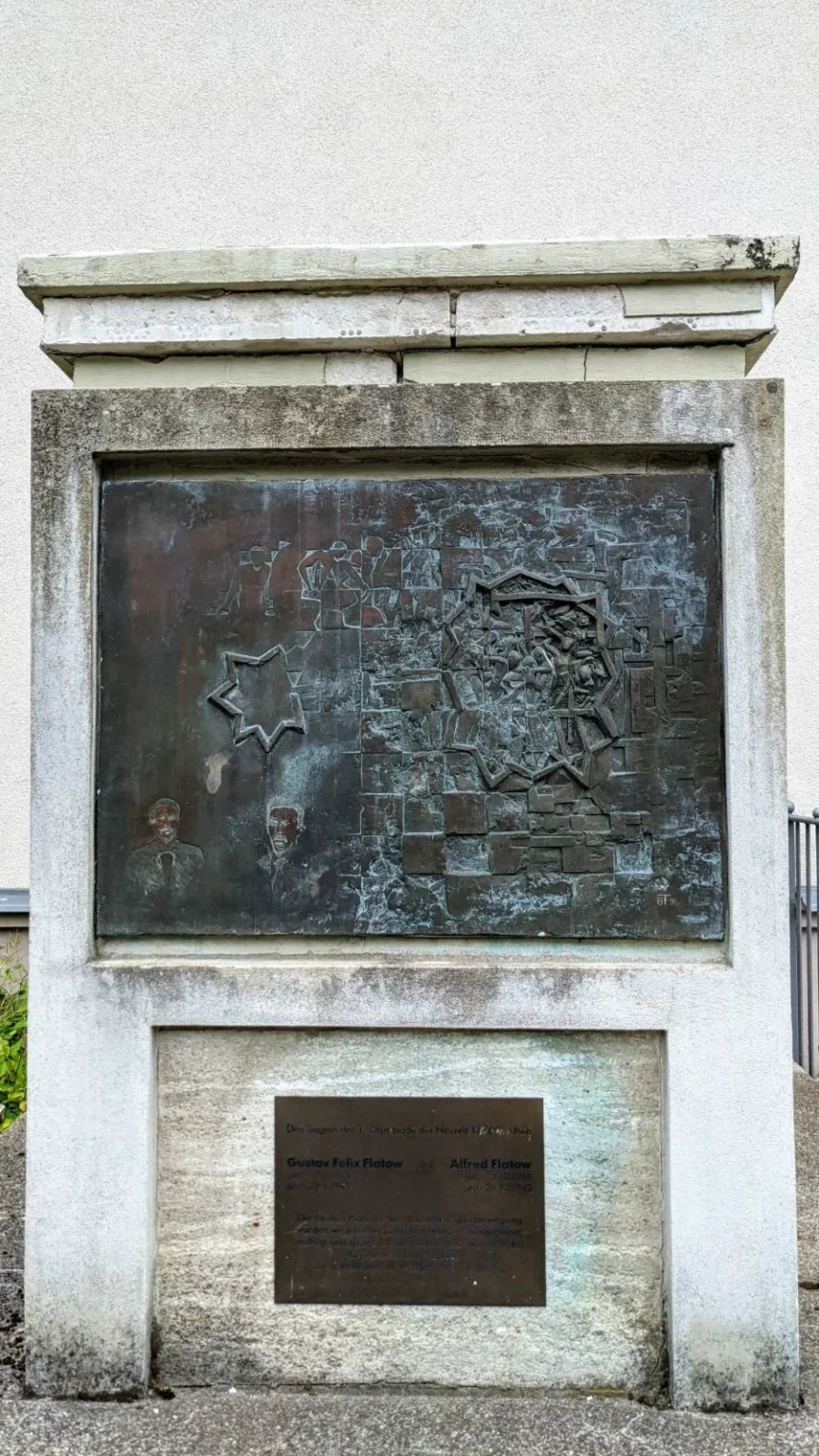 Freistehendes, hell verputztes und fleckiges Denkmal vor einer Putzwand: oben ein Bronzerelief, darunter eine kleine dunkle Texttafel für Gustav Felix und Alfred Flatow an der Birkenstraße in Köpenick