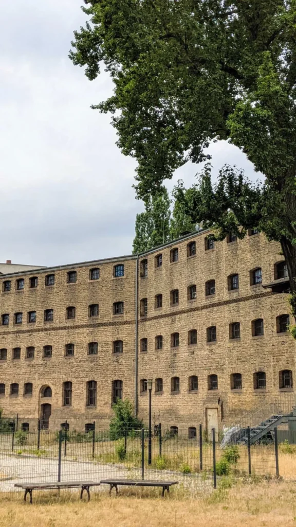 Langer Bau aus gelbem Klinker mit vielen kleinen vergitterten Rundbogenfenstern hinter einem Zaun, das frühere Amtsgerichtsgefängnis in Köpenick