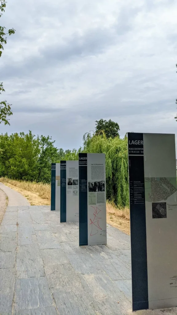 Vier hohe Informationsstelen mit der Aufschrift Lager Kaulsdorfer Straße 90 an einem gepflasterten Weg vor einer Trauerweide