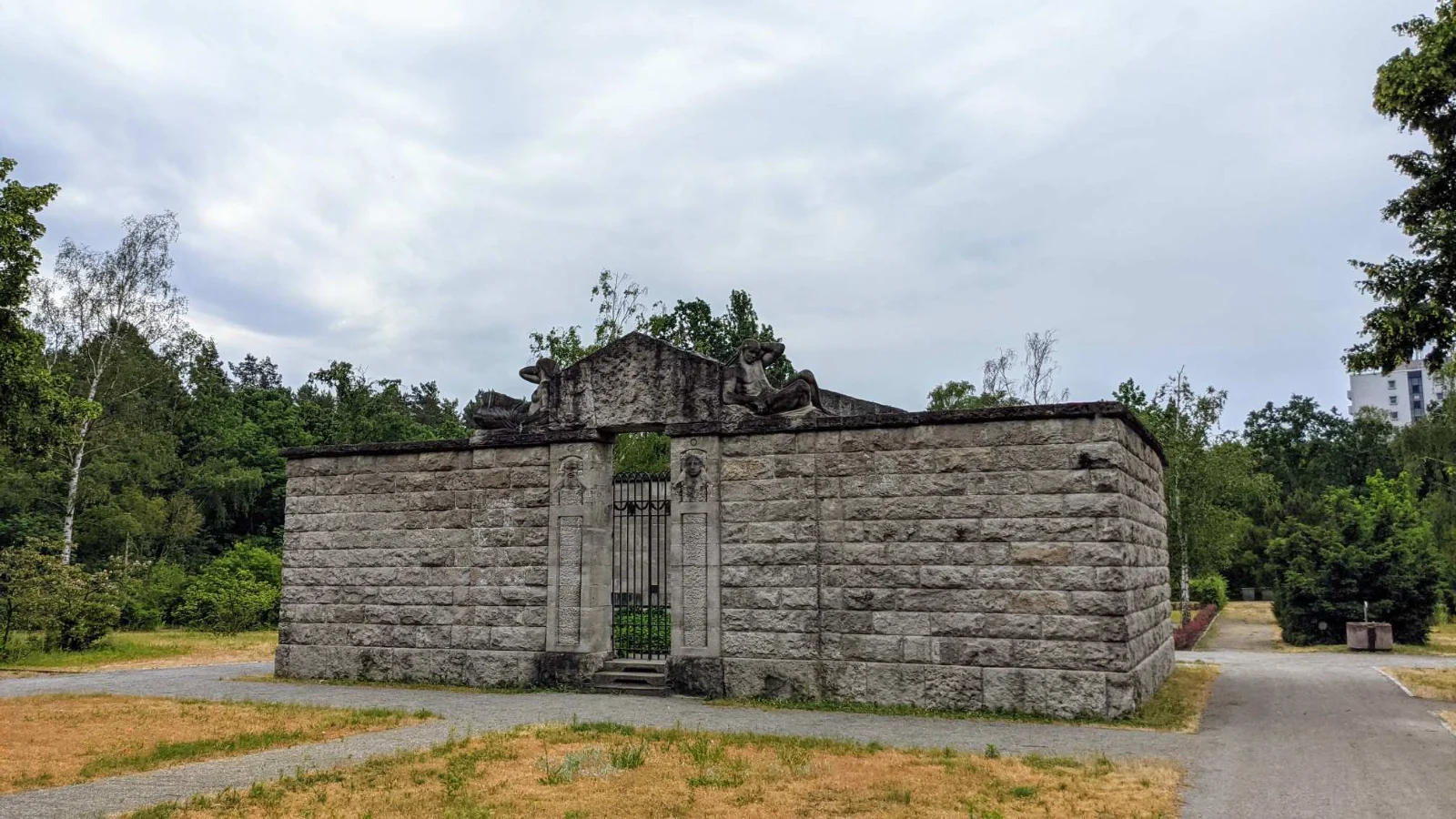 Ummauerte Grabanlage aus grauem Muschelkalk im Waldfriedhof Oberschöneweide mit Portal, schmiedeeisernem Gittertor und liegenden Steinfiguren auf dem Mauersims, umgeben von Kiefern und Birken