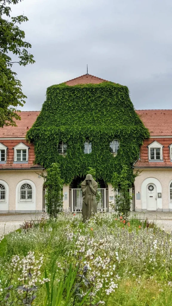 Torhaus des Alten Friedhofs Baumschulenweg mit rotem Ziegeldach und bewachsenem Mittelteil, davor die Steinfigur einer verhüllten Trauernden zwischen Blumenbeeten
