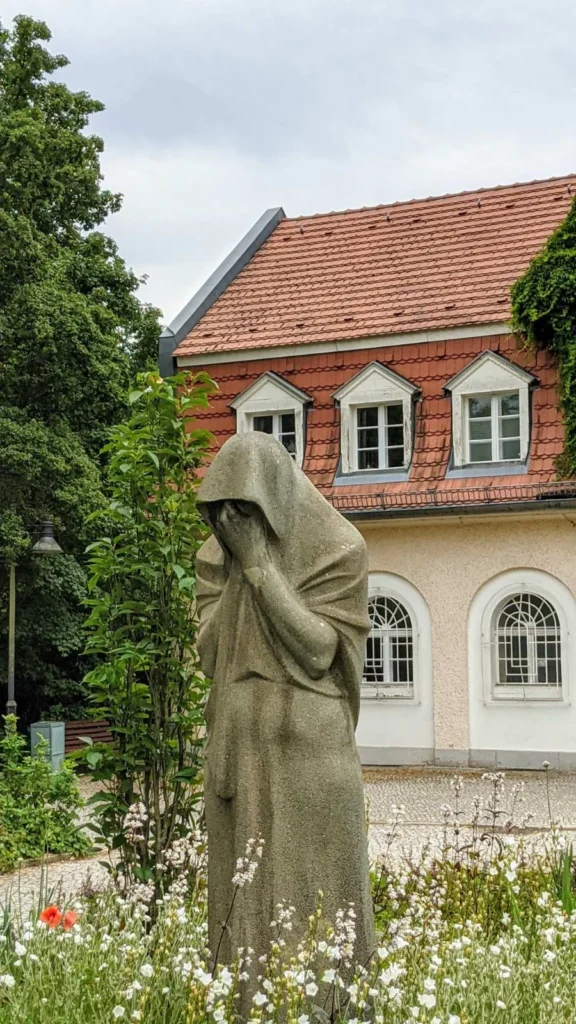 Die Steinfigur der Trauernden von Fritz Cremer von der Seite, dahinter der Seitenflügel des Torhauses, im Vordergrund weiße Blüten und roter Mohn