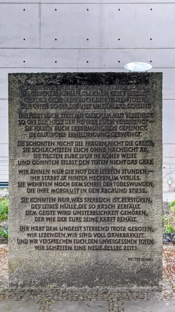 Rückseite des Gedenksteins mit einem Gedicht in sechs Strophen, dahinter die Sichtbetonwand des neuen Krematoriums