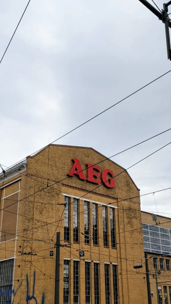 Giebel einer Industriehalle aus gelbem Klinker mit rotem AEG-Schriftzug und hohen Fenstern, davor Oberleitungen