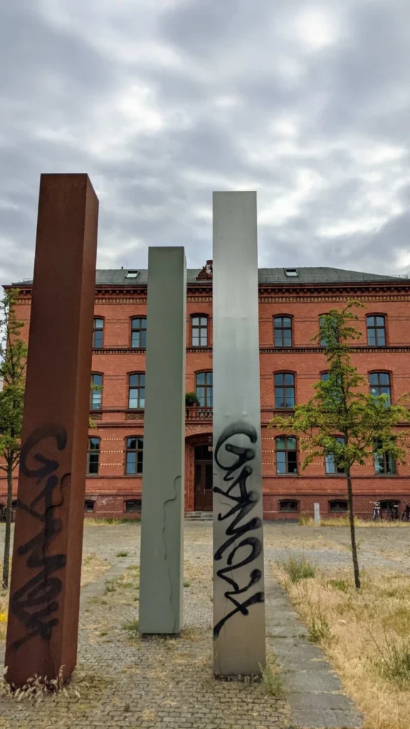 Drei hohe Vierkantstelen auf einem gepflasterten Platz am Gedenkort Rummelsburg, links rostfarben, die anderen grau, dahinter ein roter Backsteinbau