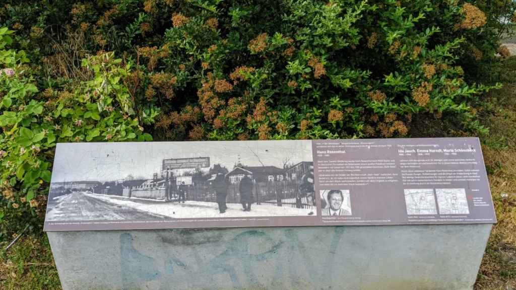 Liegende Informationstafel auf einem Betonsockel vor einer Hecke mit historischem Foto der Laubenkolonie Dreieinigkeit, zwei Textspalten, Porträt und Kartenausschnitten