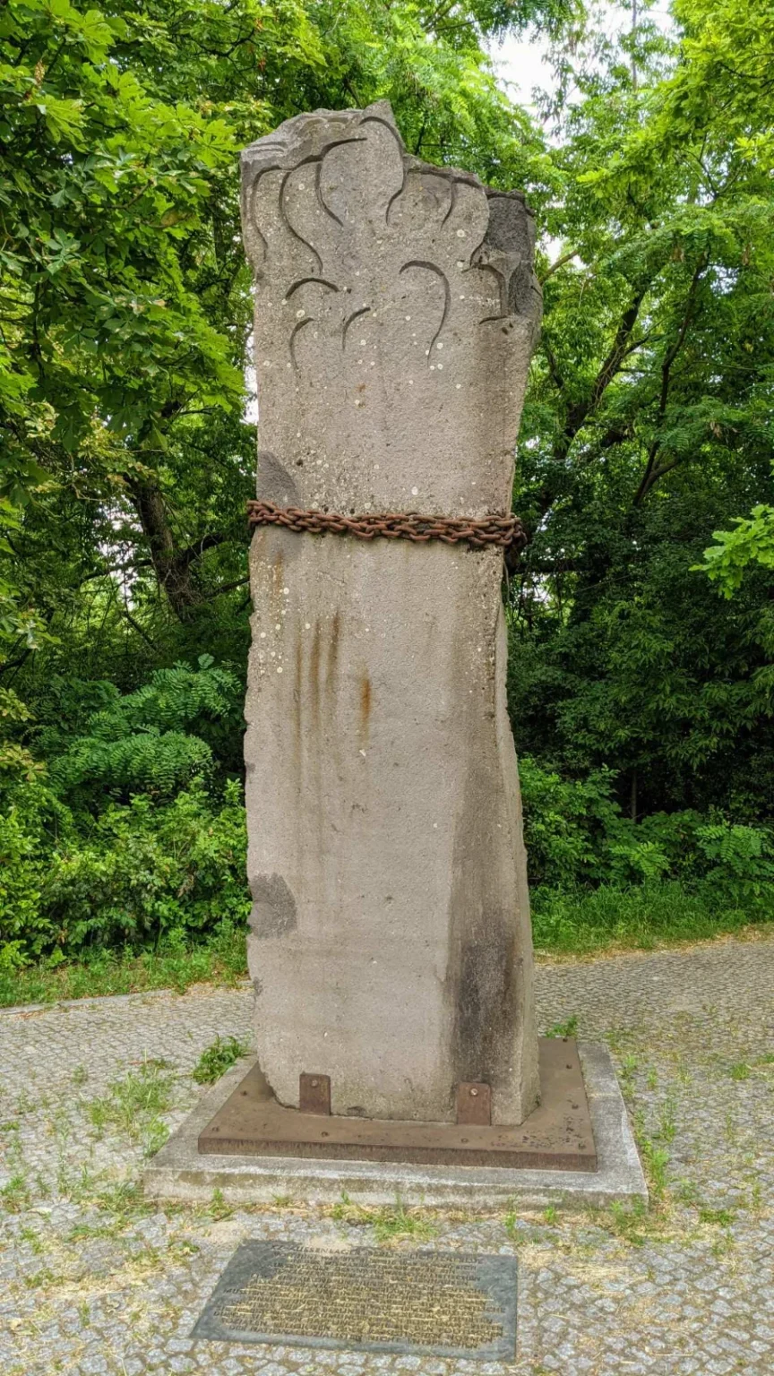 Hohe graue Steinsäule mit eingeritzten bogenförmigen Linien an der Spitze, in der Mitte von einer rostigen Eisenkette umschlungen, davor im Pflaster eine dunkle Bodenplatte mit Inschrift, dahinter dichtes Grün