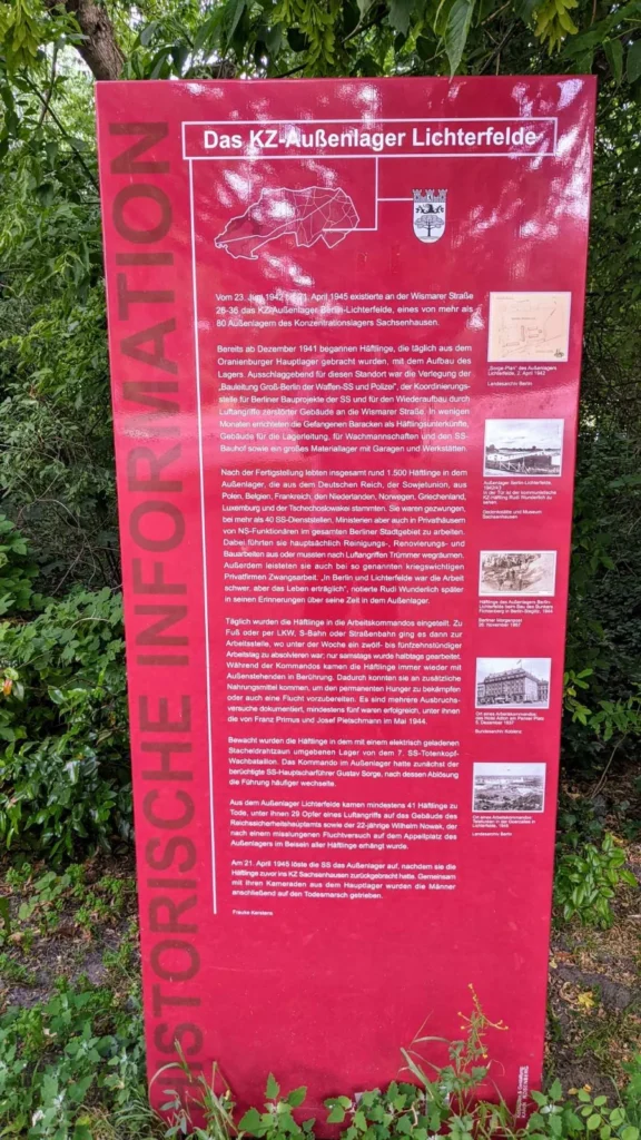 Rote Informationsstele im Gebüsch mit dem senkrechten Schriftzug Historische Information, der Überschrift Das KZ-Außenlager Lichterfelde, langem Text und fünf kleinen historischen Abbildungen