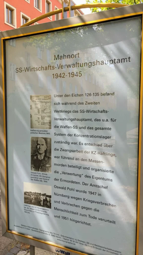 Plakatvitrine mit gelbem Rahmen, schräg von unten fotografiert, mit dem Titel Mahnort SS-Wirtschafts-Verwaltungshauptamt 1942 bis 1945, einem Text und drei historischen Abbildungen, im Glas spiegeln sich Bäume und eine Fassade