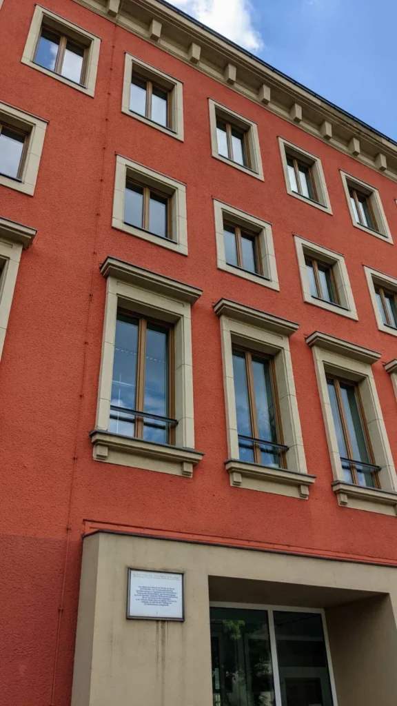 Rot verputzte Fassade mit hellen Sandsteinrahmungen und Konsolgesims von schräg unten, unten ein heller Eingangsvorbau mit einer weißen Berliner Gedenktafel neben der Glastür, darüber blauer Himmel