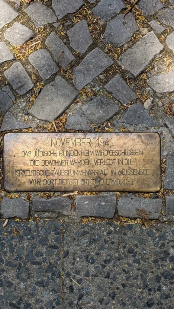 Längliche Messingplatte im Kopfsteinpflaster mit der Inschrift zur Schließung des Jüdischen Blindenheims im November 1941, in den Fugen gelbe Blütenreste