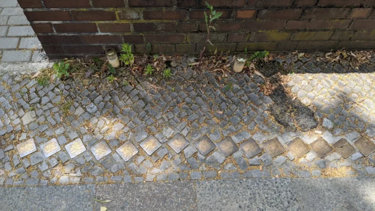 Reihe von 15 Stolpersteinen im Mosaikpflaster vor einer dunkelroten Ziegelmauer mit zwei weißen Grablichtern