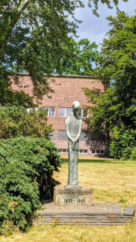 Schlanke grün patinierte Bronzefigur mit gesenktem Kopf und vor dem Körper gehaltenen Händen auf einem Steinsockel mit der Inschrift Den Verfolgten von 1933 bis 1945, dahinter eine trockene Wiese, ein roter Ziegelbau und Bäume