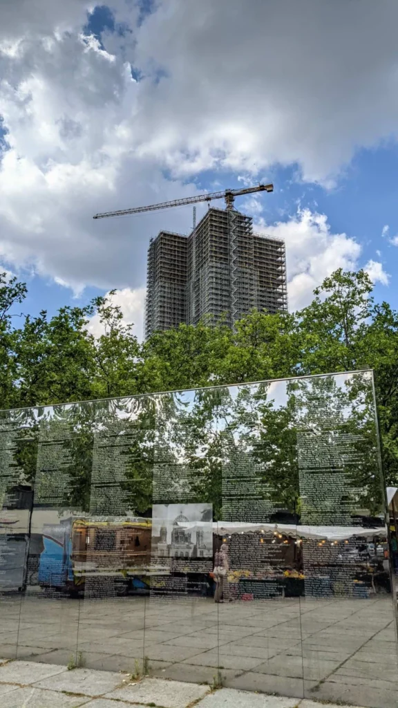 Spiegelwand mit weißen Namenslisten im unteren Bilddrittel, darin gespiegelt Marktstände mit Lichterkette, darüber Baumkronen und ein eingerüstetes Hochhaus mit Kran vor Wolkenhimmel