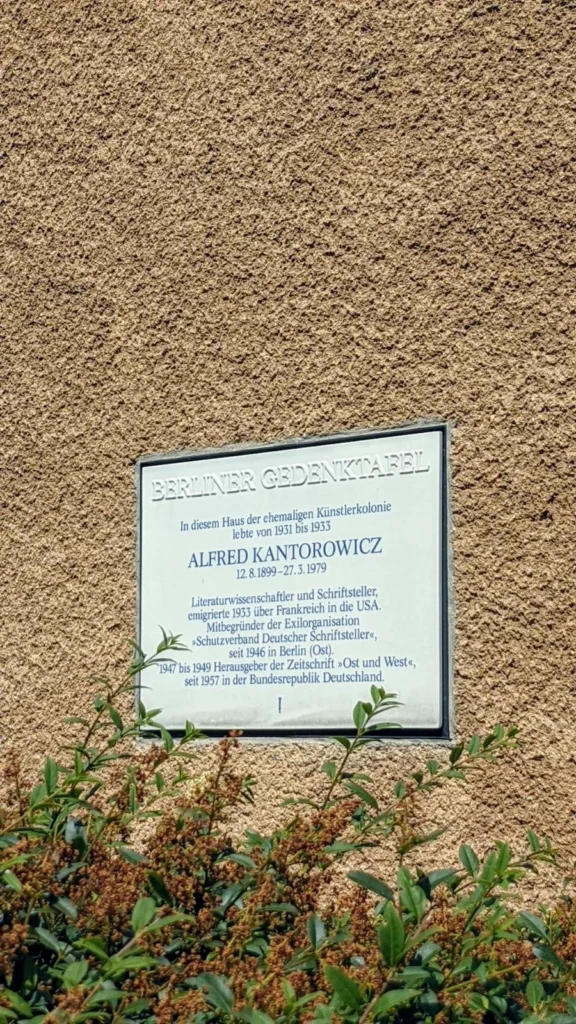 Berliner Gedenktafel für Alfred Kantorowicz an einer ockerfarbenen Rauputzwand der Künstlerkolonie, unten ragt ein Strauch ins Bild