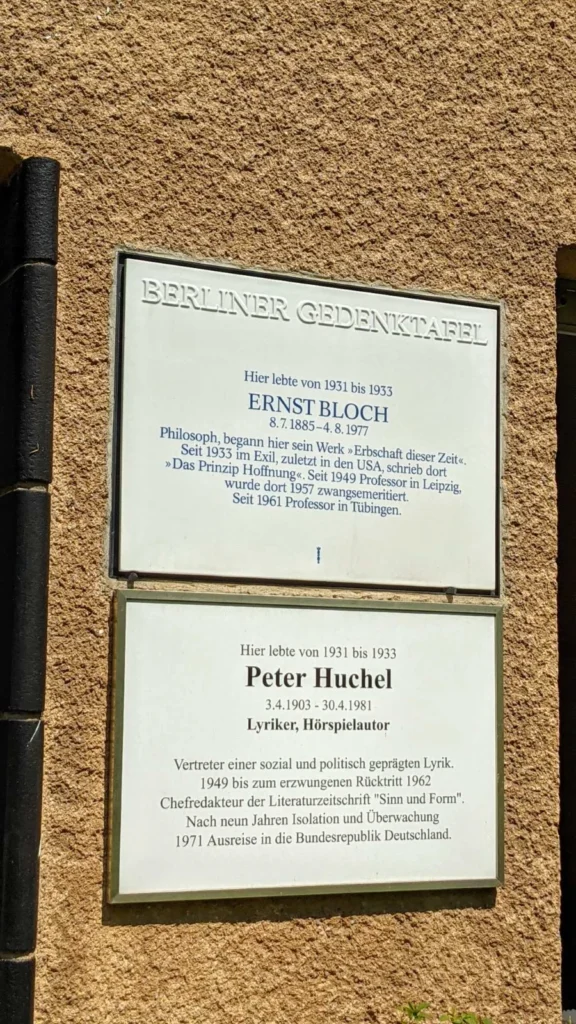 Zwei Tafeln übereinander an einer Rauputzwand: oben die Berliner Gedenktafel für Ernst Bloch, darunter eine weiße Tafel im grünen Rahmen für Peter Huchel, links ein schwarzes Fallrohr