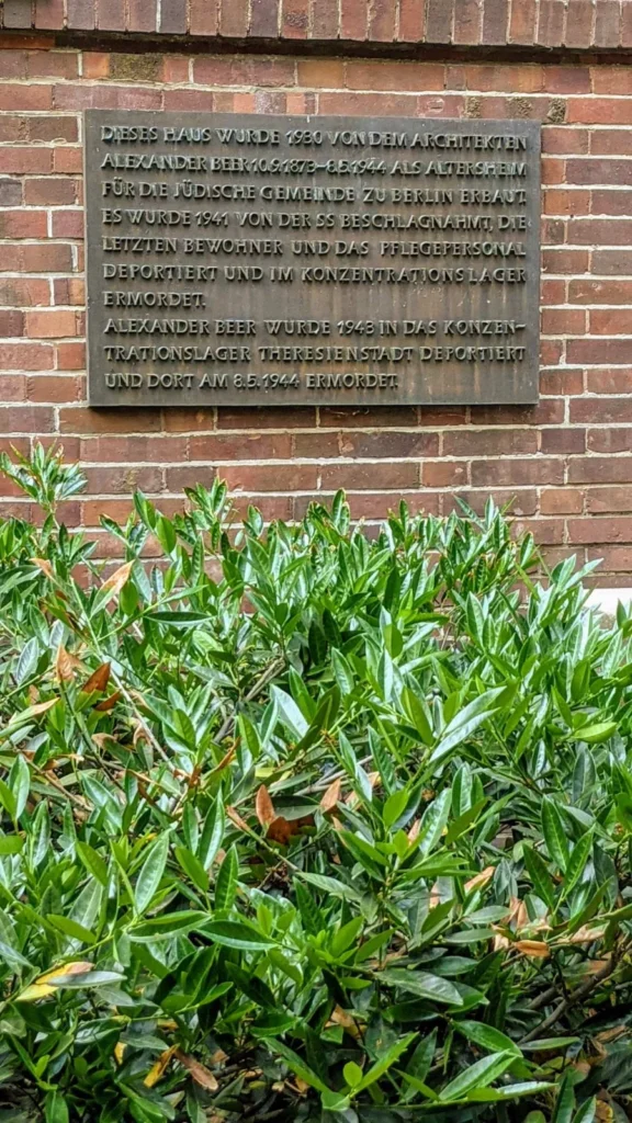 Bronzetafel an einer roten Ziegelwand, die untere Bildhälfte ist von einer Kirschlorbeerhecke ausgefüllt, sie erinnert an das Altersheim der Jüdischen Gemeinde, das Alexander Beer gebaut hat