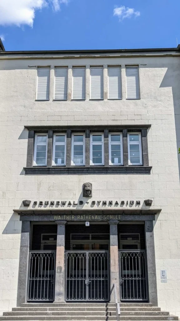 Frontale Ansicht eines Schulportals aus heller Natursteinfassade mit drei vergitterten Türöffnungen und Freitreppe, darüber die Schriftzüge Grunewald-Gymnasium und Walther Rathenau-Schule und ein Kopfrelief mit Helm