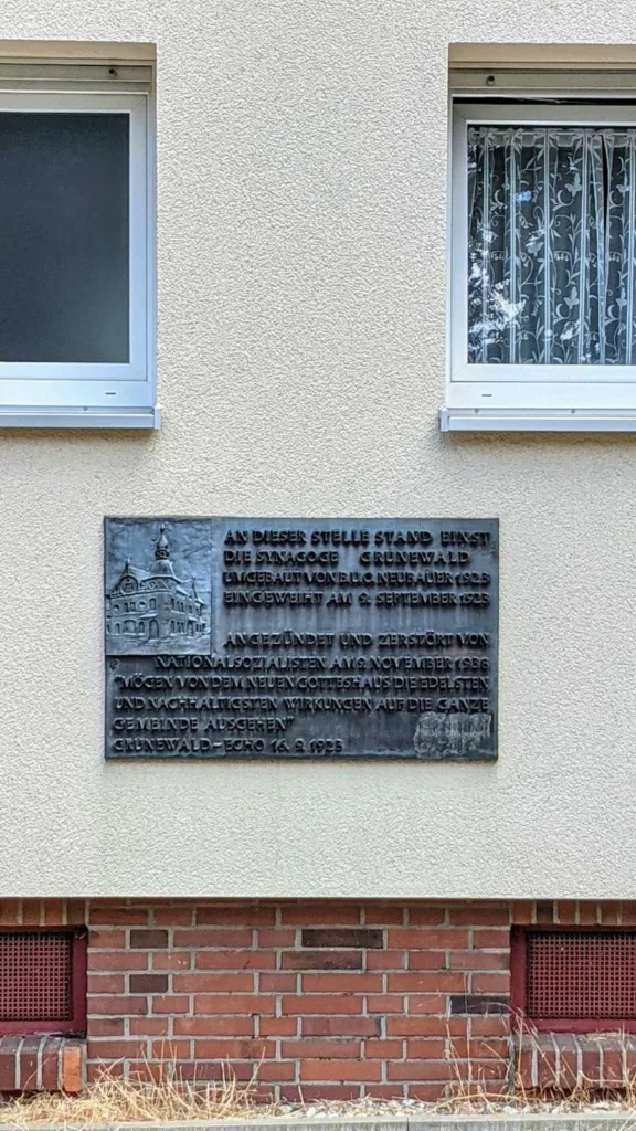 Bronzetafel an einer hell verputzten Hauswand zwischen zwei Fenstern, links mit dem Relief eines Gebäudes mit Turm, sie erinnert an die Synagoge Grunewald