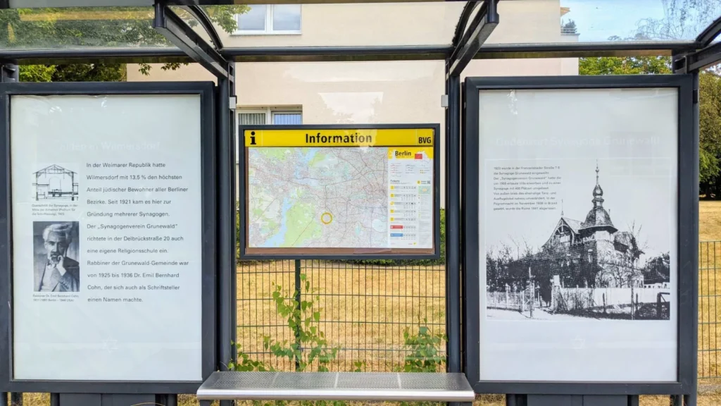 Wartehalle einer Bushaltestelle mit zwei Plakatvitrinen und einer gelben Informationstafel der BVG dazwischen, das linke Plakat trägt den Titel Juden in Wilmersdorf, das rechte Gedenkort Synagoge Grunewald, beide mit einem Davidstern