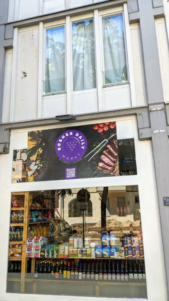 Schaufenster eines Lebensmittelgeschäfts mit dem Schild Kosher Daily Markt, in der Auslage Snacks und eine lange Reihe Weinflaschen, darüber ein Obergeschoss mit drei Fenstern
