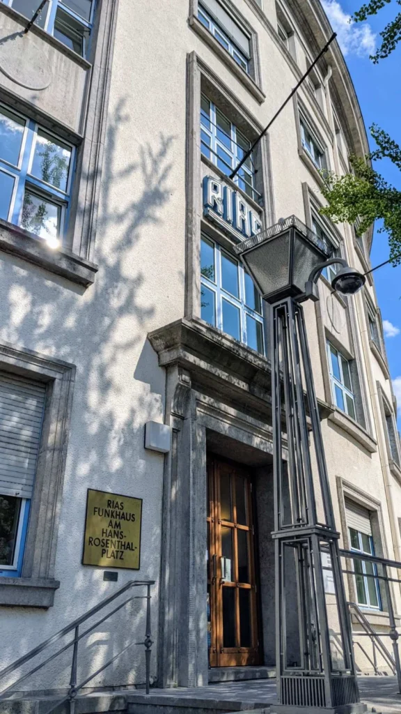 Eingang eines geschwungenen Putzbaus von schräg unten, an der Fassade ein blau-weißer Neonschriftzug RIAS, daneben ein Messingschild, im Vordergrund ein Laternenmast mit Kamera