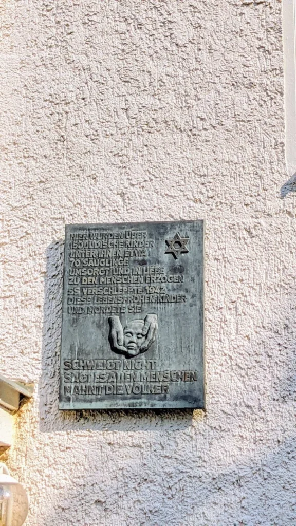 Bronzene Gedenktafel mit Davidstern und zwei Händen, die ein Kindergesicht halten, an einer hellen Hauswand des früheren jüdischen Kinderheims in Niederschönhausen
