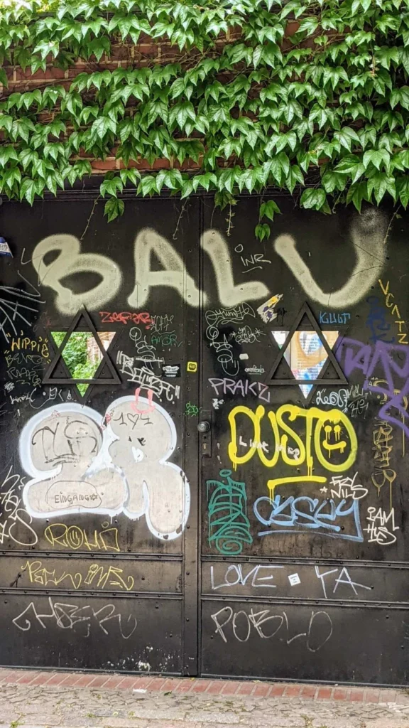 Schwarzes Stahltor mit zwei Davidstern-Fenstern, dicht mit Graffiti bedeckt, am Zugang zum Judengang am Kollwitzplatz